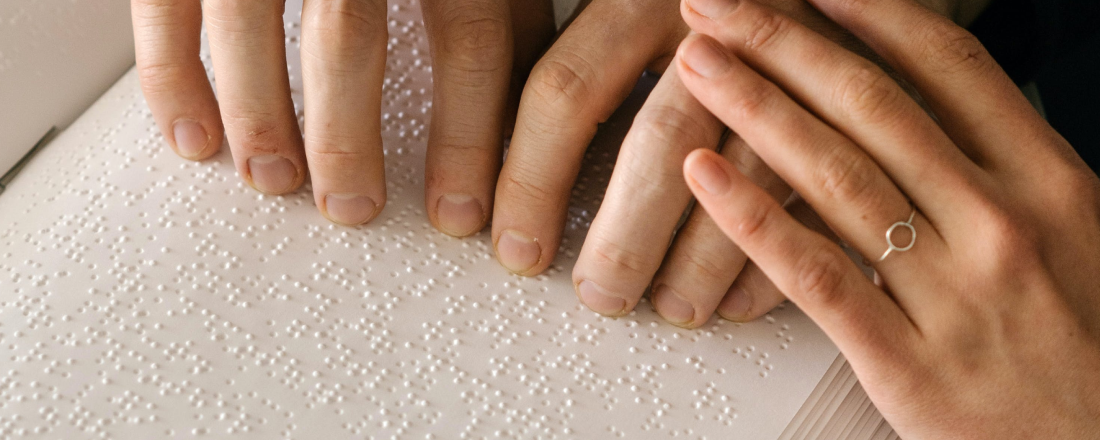 Zbliżenie na 3 ręce. Dwie czytają Pismo Braille’a, a trzecia je kieruje po tekście.