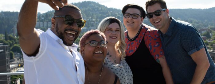 Grupa znajomych robi sobie zdjęcie z ręki na tle gór. Na zdjęciu jest pięć różnorodnych osób, które reprezentują inkluzywność.