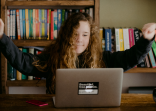 Dziewczyna z uniesionymi rękami w górę patrzy na ekran laptopa przed nią. W tle półka z książkami. Zdjęcie nawiązuje do WCAG 3.1.1 Język strony.